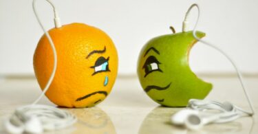 Imagem de uma laranha e uma maçã verde. Uma está bonita e a outra já está mordida. Ambas usam um fone de ouvido e estão representando o sentimento de inveja.