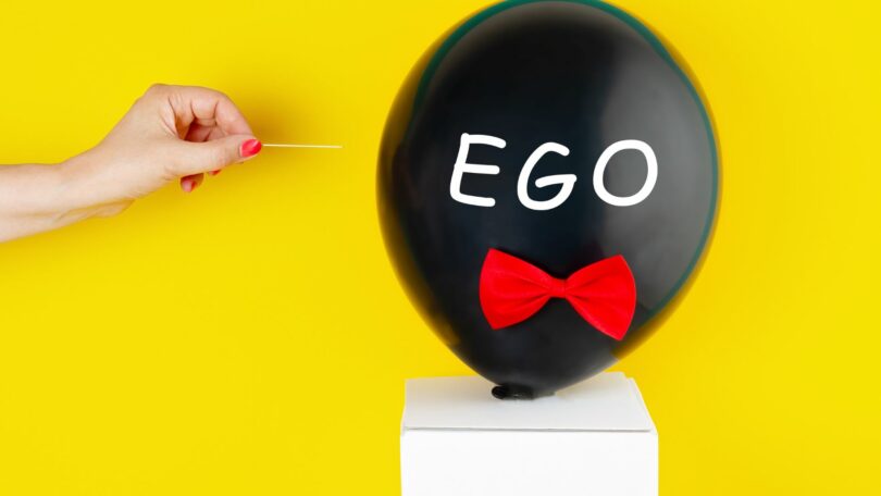 Imagem de fundo amarelo e em destaque um balão preto com o texto Ego escrito sobre ele e mão de uma mulher que vai estourar o balão com agulha. A imagem traz os conceitos de orgulho e egoísmo.