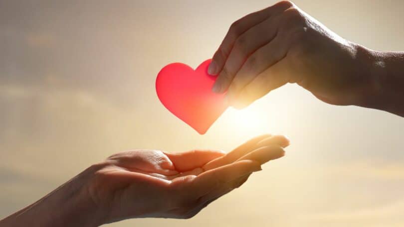Imagem com o pôr do Sol ao fundo e em destaque uma duas mãos, sendo que uma delas segura um coração vermelho.