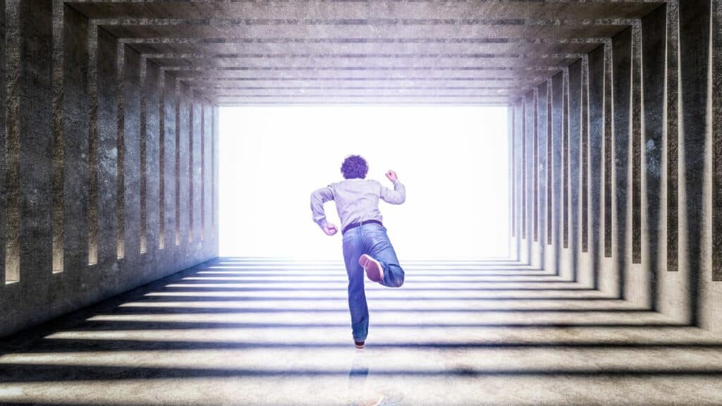 Imagem de um corredor com uma luz ao fundo. Em destaque, um homem usando calça jeans, camisa cinza e tênis, correndo em busca de encontrar uma saída.

