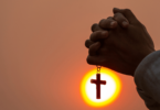 Mãos juntas, segurando uma corrente com cruz, em sinal de oração. O fundo está iluminado de amarelo e laranja.