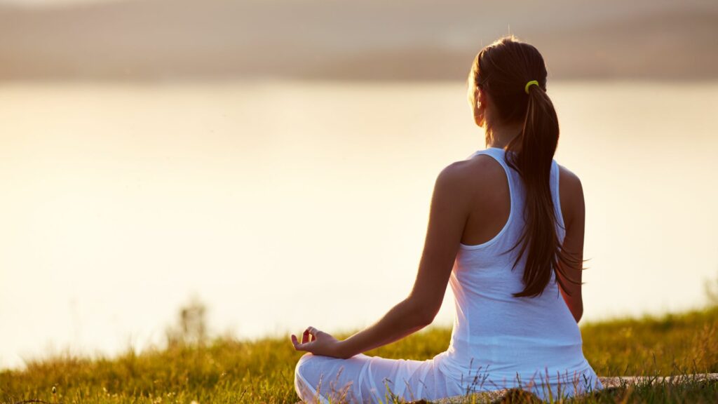 Imagem de uma mulher usando uma roupa branca, sentada em um gramado em frente a um lago fazendo meditação.
