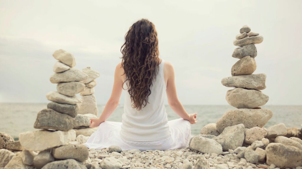 Imagem de uma mulher sentada, olhando para o mar. Ao lado dela, várias pedras. Ela está buscando o equilíbrio fazendo uma meditação.

