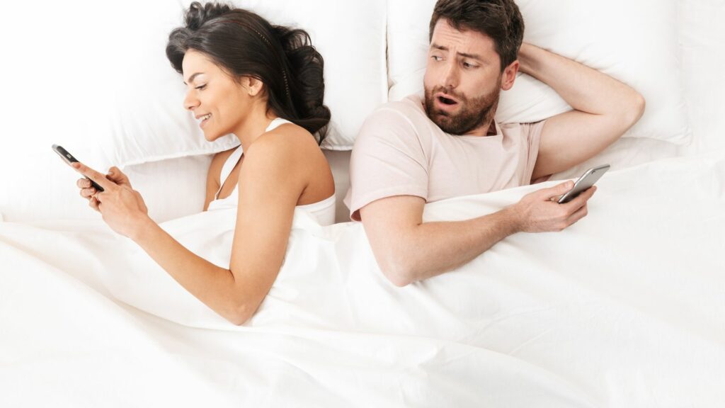 
Imagem de um casal deitado sobre uma cama. Eles estão de costas um para o outro e ambos estão mexendo e conversando no celular, representando a infidelidade.
