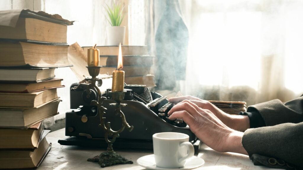 Imagem de uma mesa e sobre ela uma máquina de escrever antiga, várfios livros, um castiçal com velas acesas, uma xícara de café. Em destaque, mãos de um homem escrevendo um texto na máquina.