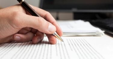 Imagem da mão de um homem, segurando uma caneta preta. Ele está escrevendo várias frases em um papel sobre uma mesa.