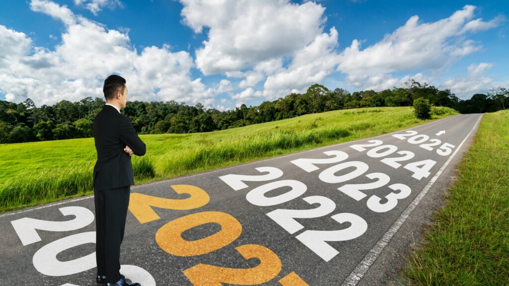 Imagem de uma estrada e nela está escrito em letras garrafais os anos de 2020 a 2025 e uma seta mostrando o fim da estrada. Em destaque um homem usando um terno preto, em pé nesta estrada olhando para o futuro.
