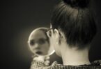 Imagem de fundo preto e em destaque uma mulher com cabelo preso e óculos, olha em um espelho, trazendo o conceito de autocrítica.