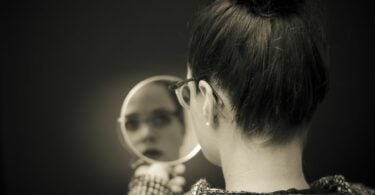 Imagem de fundo preto e em destaque uma mulher com cabelo preso e óculos, olha em um espelho, trazendo o conceito de autocrítica.