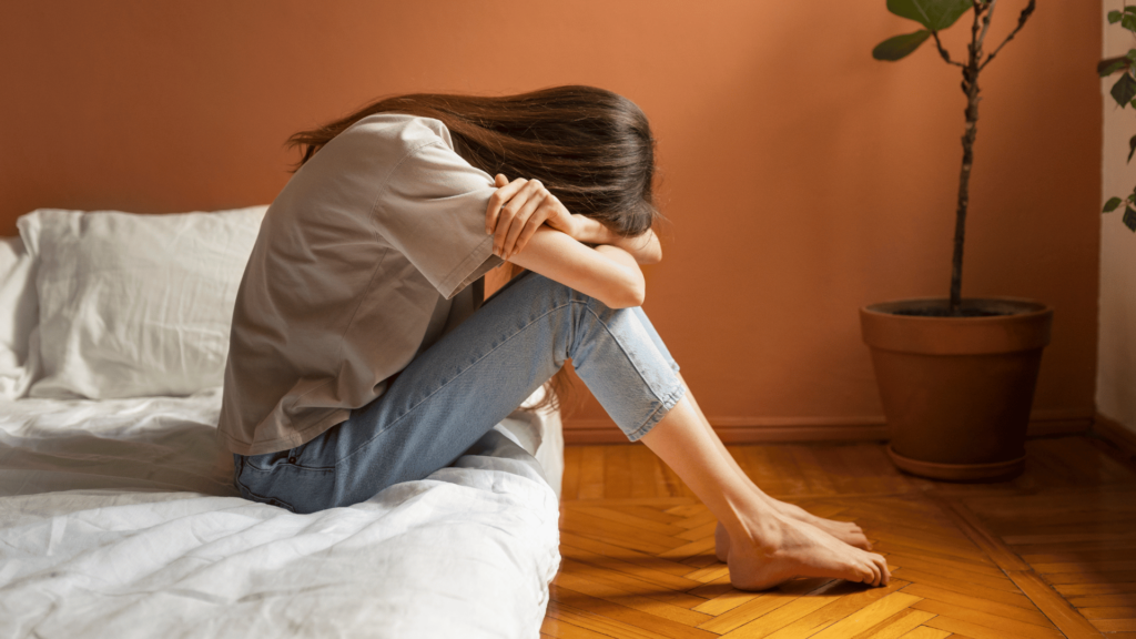 Mulher sentada na cama, com a cabeça e os braços debruçados sobre as pernas, com sentimento de baixa-autoestima.