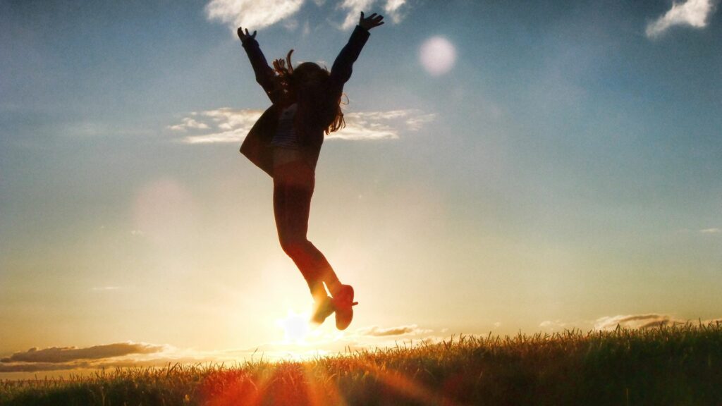 Imagem de um lindo pôr do sol em um campo. Em destaque a silhueta de uma mulher feliz e motivada, dando um salto para a vida.
