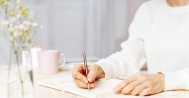 Imagem de uma mulher usando um moletom branco. Ela está sentada e sobre uma mesa, está escrevendo em um carderno. Ao lado, uma caneca de chá e um arranjo floral.