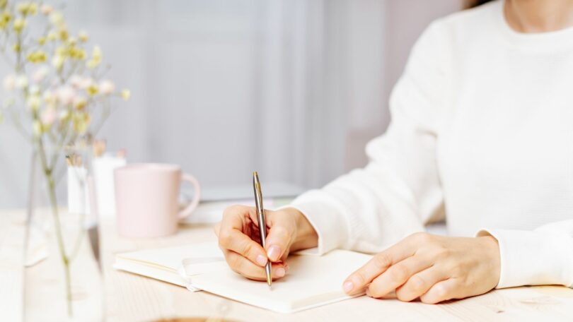 Imagem de uma mulher usando um moletom branco. Ela está sentada e sobre uma mesa, está escrevendo em um carderno. Ao lado, uma caneca de chá e um arranjo floral.