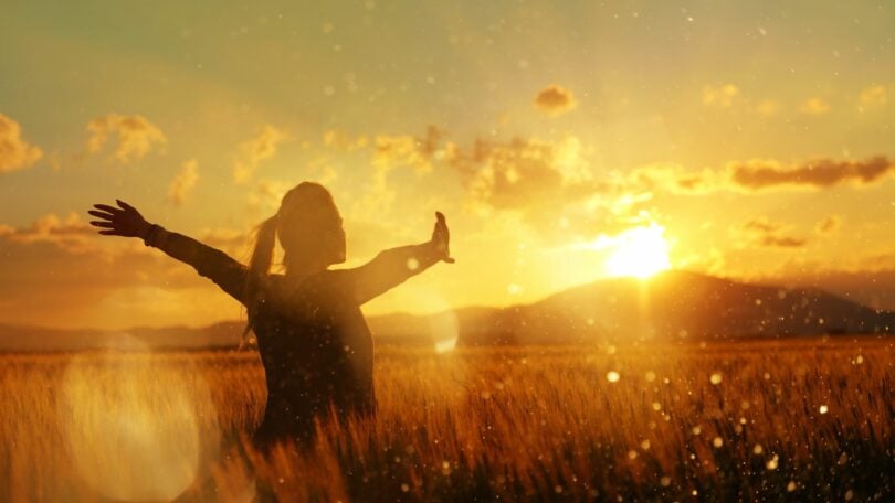 Imagem de um lindo campo de trigo e ao fundo um pôr do Sol. Em destaque, uma mulher de braços abertos, de frente para o Sol, trazendo o conceito de cura das dores da alma.