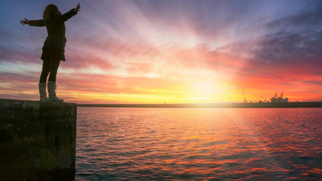 Imagem de um lindo mar e ao fundo o pôr do sol. Em destaque, uma mulher sobre uma pedra, em pé e de braços abertos, transmitindo o conceito de viver plenamente o hoje, de ser você mesma.
