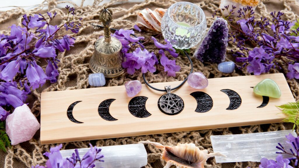 Imagem de uma decoração feita com pedras de quartzo roxo para o altar pagão da bruxa da lua cheia feito com imagens da com fases da lua, cristais, flores roxas e pingente de pentagrama.