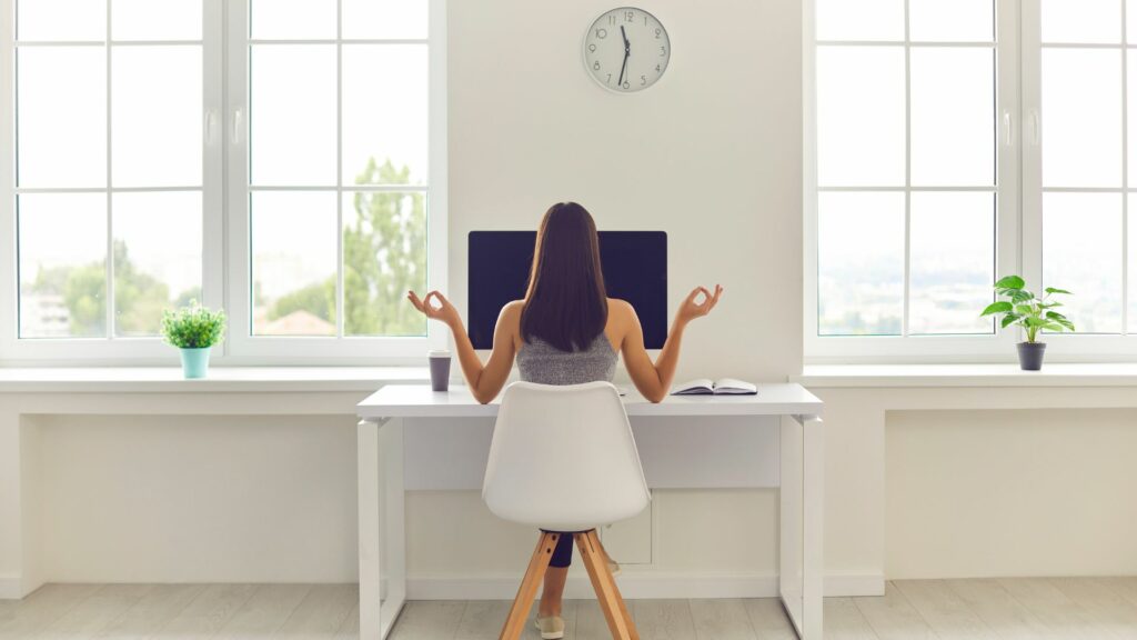 Imagem de uma mulher de costas, fazendo uma pausa no trabalho e meditando. Ela está sentada na mesa do escritório com um computador e ao lado, um copo com café, trazendo o conceito de Mindfulness.