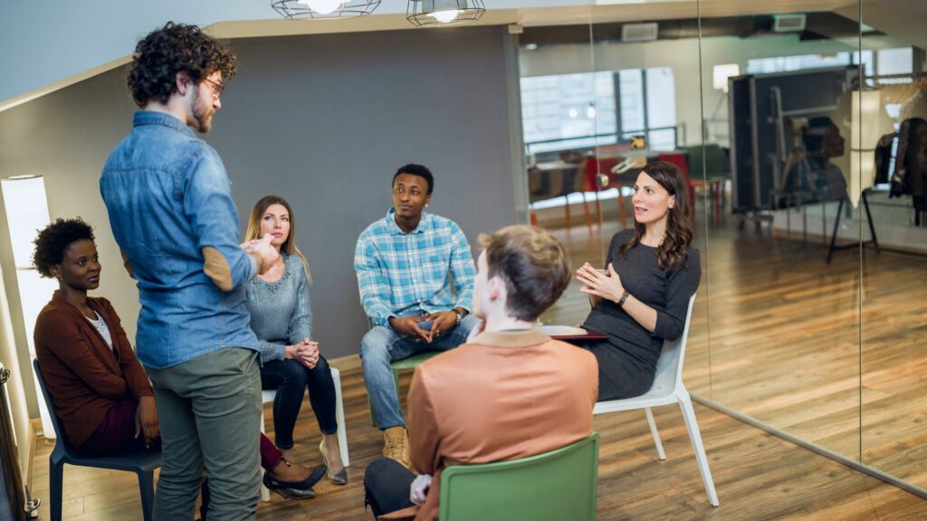 Imagem de várias pessoas sentadas, participando de um workshop em grupo. Em pé, um jovem, ministrando uma pequena palestra para eles sobre resolução de problemas.
