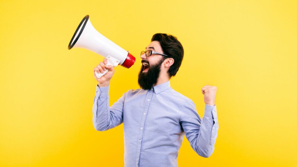 Imagem de fundo amarelo e em destaque um homem de barba, vestindo uma camisa azul claro e usando óculos. Ele está segurando um megafone em uma de suas mãos, trazendo o conceito de estar gritando.
