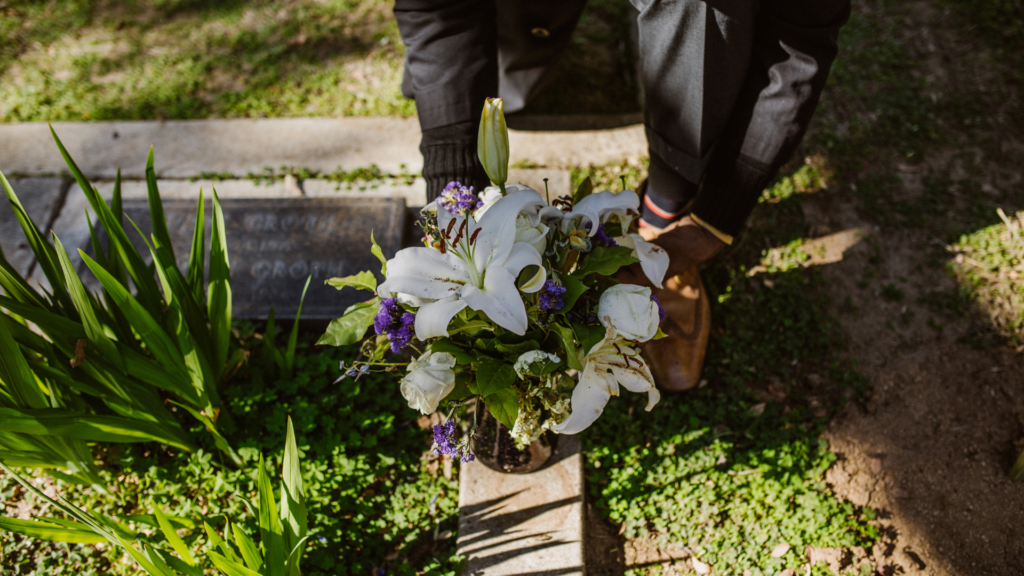 Imagem de uma pessoa usando uma roupa preta. Ela está colocando flores em um túmulo.