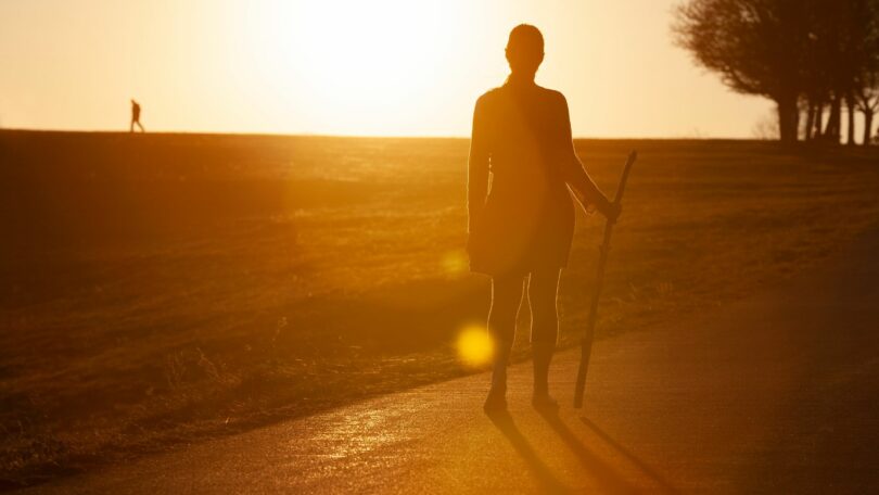 Imagem de uma mulher fazendo romaria em uma estrada de terra em pleno pôr do Sol.