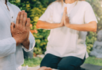 Duas pessoas sentadas em tapete sobre o ar livre, um jardim. Elas estão em posição de meditação, com as mãos juntas.