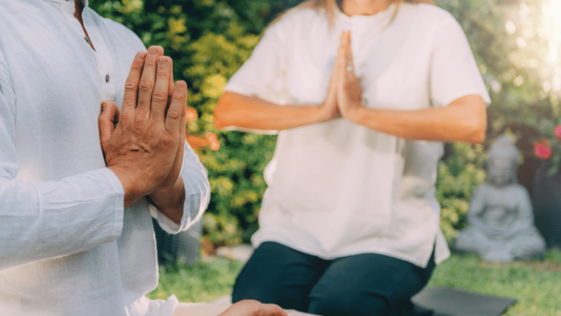 Duas pessoas sentadas em tapete sobre o ar livre, um jardim. Elas estão em posição de meditação, com as mãos juntas.