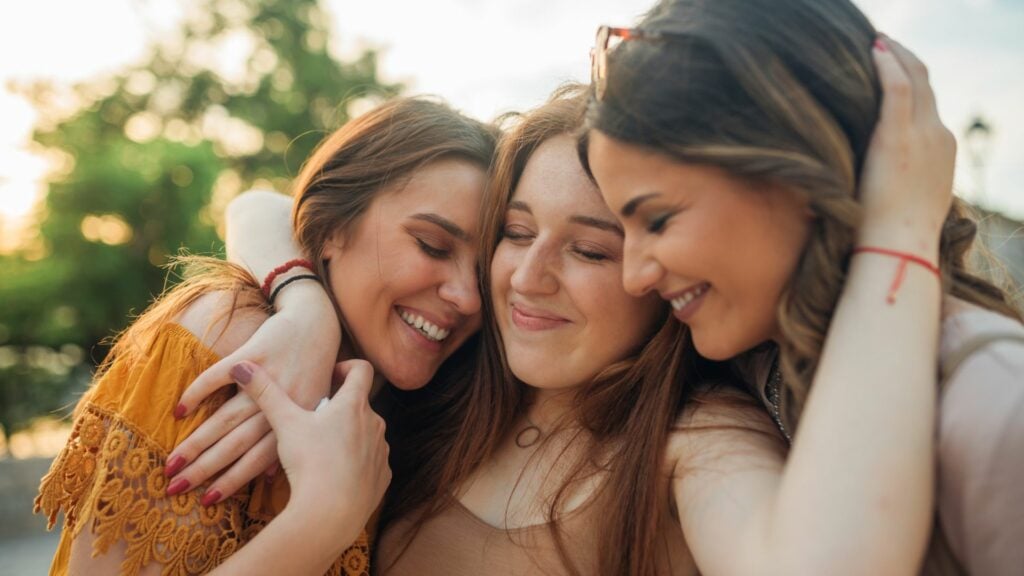 Imagem com três mulheres se abraçando, sorrindo, olhando para baixo e demonstrando apoio à mulher que está no meio das outras duas localizadas nas extremidades.