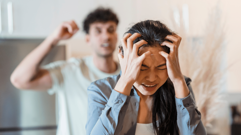 Um casal está tendo uma discussão. A mulher de aparência asiática está no centro da imagem com as duas mãos na cabeça e a expressão de frustração ou desgaste. Atrás dela, há um homem desfocado com o braço direito levantado, discutindo e com a expressão de raiva.