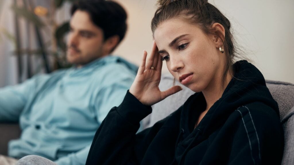 Um casal composto por um homem e uma mulher estão sentados em um sofá cinza. A mulher está em destaque na imagem com a mão na cabeça e a feição do rosto aflita. O homem está ao lado da mulher na imagem, desfocado e olhando para frente.