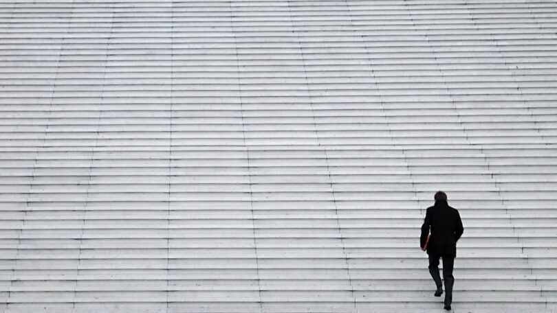 Um homem de roupa preta e com uma pasta vermelha na mão sobe sozinho uma grande escadaria branca. Ele está posicionado no cantor inferior esquerdo da imagem. A maior parte da foto é composta pela escadaria que, em contraste com o homem subindo e ocupando um pequeno espaço da imagem, dá a sensação de solidão e vazio.