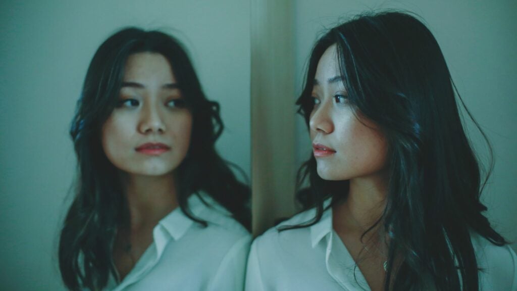 Mulher jovem de aparência asiática com cabelos pretos e lisos e que veste uma camisa branca olha para o espelho, o qual a reflete na imagem. Há um fundo verde atrás da mulher.