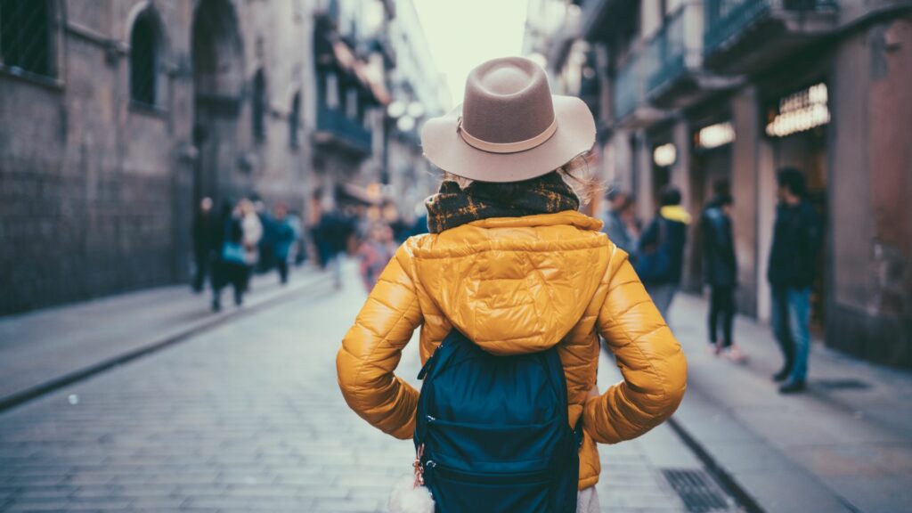 Mulher de casaco amarelo, cachecol, chapéu bege e que usa uma mochila azul caminha por uma rua. A mulher aparenta ser uma turista e está centralizada de costas na imagem. Ao fundo, há uma rua, prédios e pessoas desfocadas.