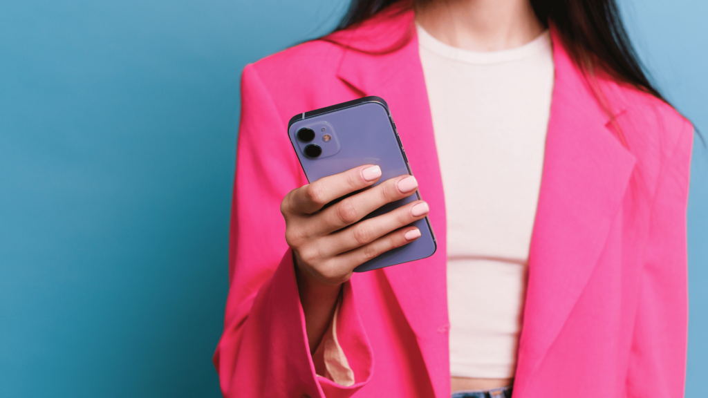 Uma mulher de camiseta branca e blazer rosa levemente desfocada segura um celular na mão direita. O celular está em foco e há um fundo azul atrás da mulher na imagem.