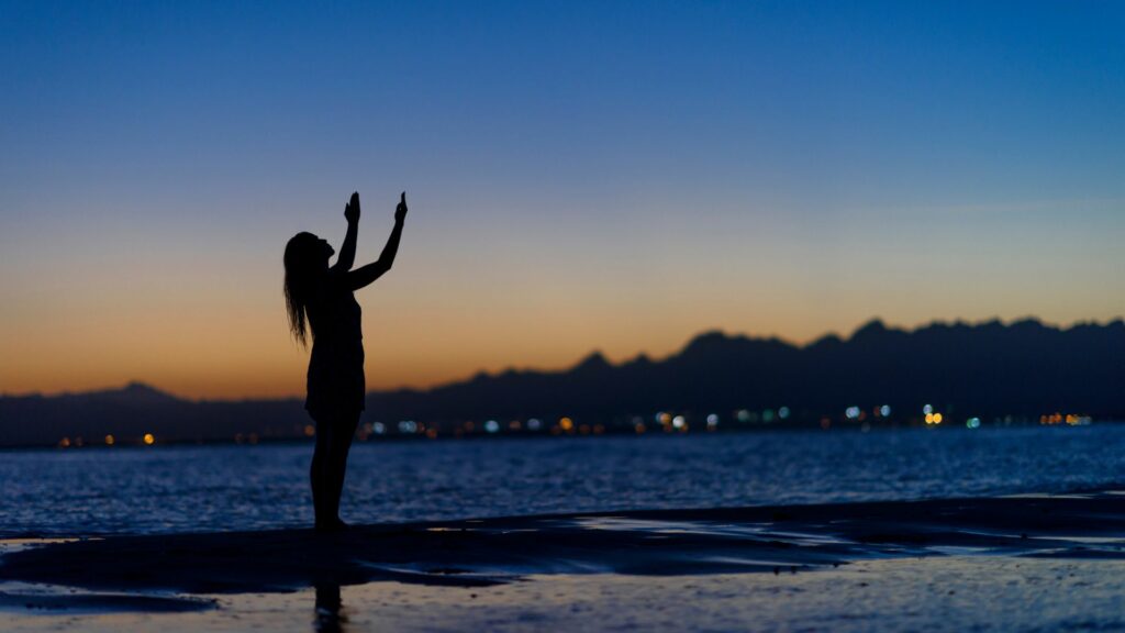 Silhueta de mulher posicionada de lado. Ela está em pé em uma praia diante do pôr do sol com os braços e o rosto levantados para cima, conceituando uma oração ou agradecimento. Ao fundo da imagem, há o mar, as luzes da cidade e as montanhas desfocadas.