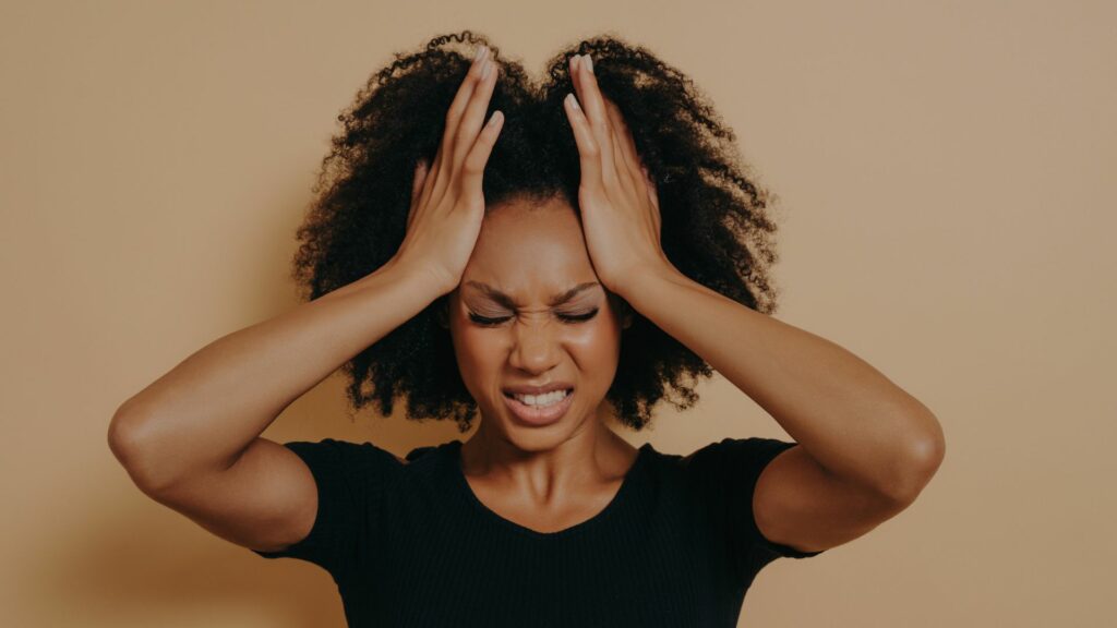 Mulher de cabelos castanhos e enrolados está com as duas mãos na cabeça e uma expressão de irritação ou frustração no rosto. Ela usa uma blusa preta. Atrás dela, há um fundo bege.