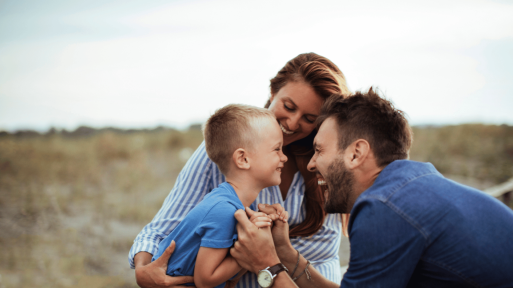 Imagem com o foco em um casal composto por uma mulher e um homem segurando e sorrindo para um menino, provavelmente seu filho. O menino está curvado e sorri. Ao fundo, há um ambiente de natureza e o céu desfocados.