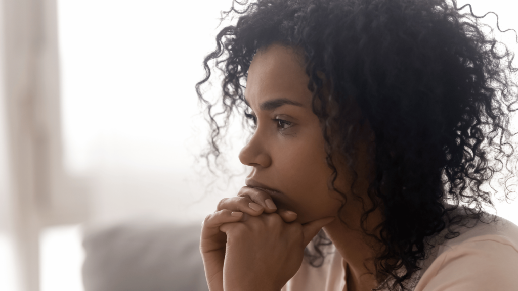 Foco no rosto de uma mulher negra de cabelos castanhos pensativa. Ela está com as duas mãos entrelaçadas, apoiando o queixo. Ao fundo, há um cômodo branco desfocado.