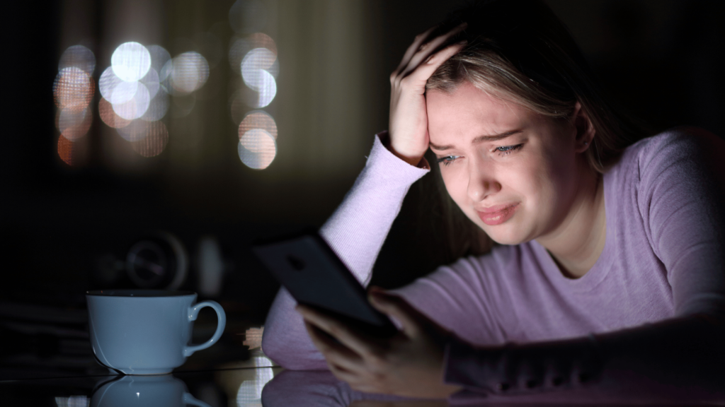 Foco em uma mulher loira que está segurando e olhando para um celular com a expressão de insatisfação. Ela está em um cômodo escuro e desfocado ao fundo e o celular ilumina o seu rosto. Ela se apoia em uma mesa e há uma xícara azul na sua frente sobre a mesa.