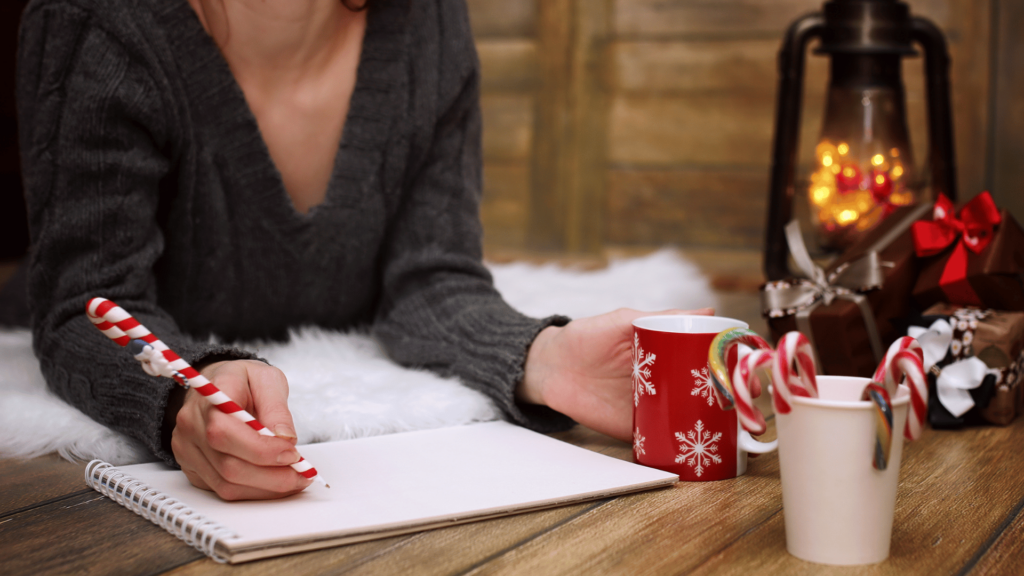 Os dois braços de uma mulher estão no foco da imagem. Ela segura uma caneta vermelha e branca enfeitada de Natal em uma mão e a apoia em um caderno branco. Ela veste uma blusa cinza e segura uma caneca vermelha na outra mão. Há decorações natalinas sobre a mesa em que ela se apoia.