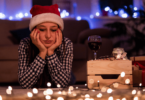 Mulher com toca de Papai Noel na cabeça está deitada no chão com as duas mãos apoiando o rosto e tem uma expressão pensativa. Ela olha para baixo e, no chão, há luzes natalinas ligadas e um celular. Ao fundo, há uma sala enfeitada para o Natal que está desfocada.