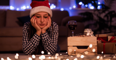 Mulher com toca de Papai Noel na cabeça está deitada no chão com as duas mãos apoiando o rosto e tem uma expressão pensativa. Ela olha para baixo e, no chão, há luzes natalinas ligadas e um celular. Ao fundo, há uma sala enfeitada para o Natal que está desfocada.