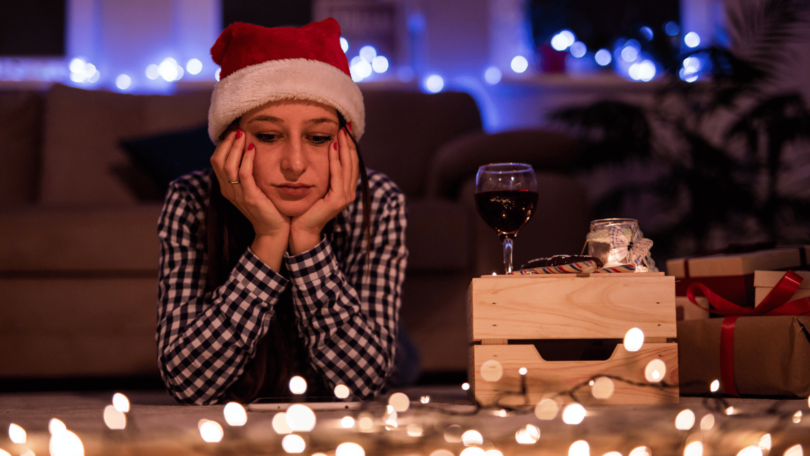 Mulher com toca de Papai Noel na cabeça está deitada no chão com as duas mãos apoiando o rosto e tem uma expressão pensativa. Ela olha para baixo e, no chão, há luzes natalinas ligadas e um celular. Ao fundo, há uma sala enfeitada para o Natal que está desfocada.