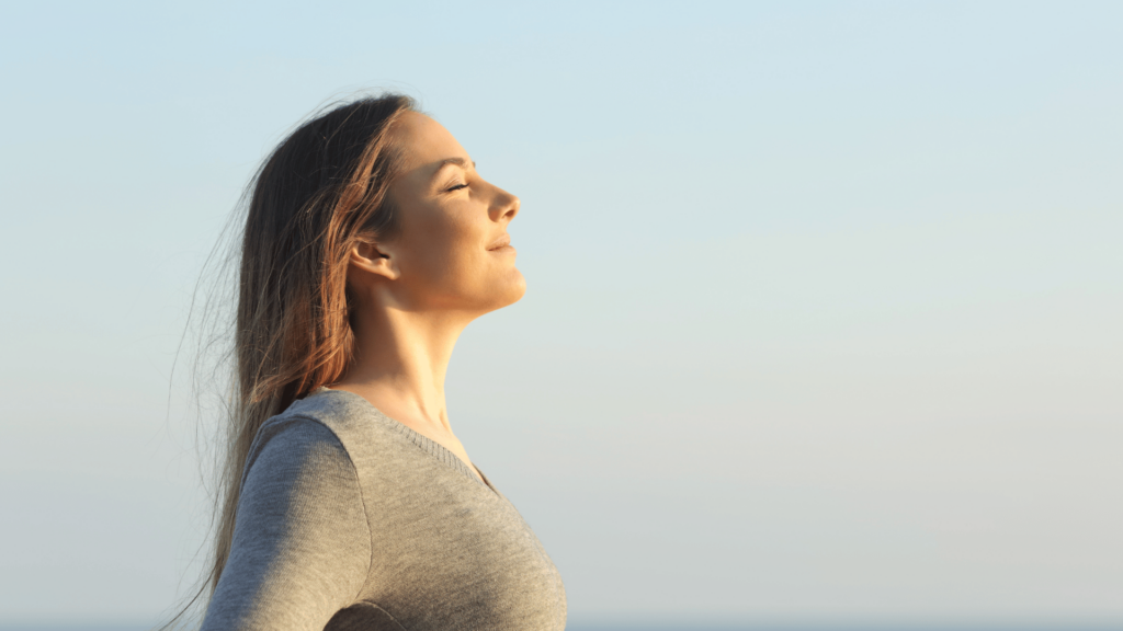 Mulher loira respira ar fresco em uma praia. Ela usa uma blusa cinza, a luz do sol bate no seu rosto, ela levanta a cabeça e respira fundo com os olhos fechados.