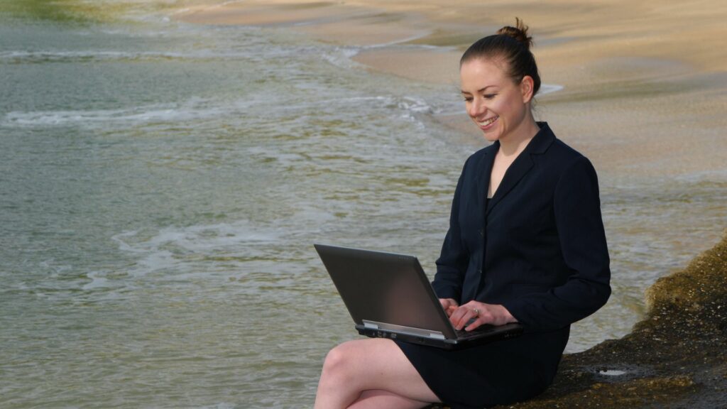 Imagem de uma mulher executiva, sentanda em uma pedra na beira do amor, ela está usando o notebook e trabalhando. A imagem traz o conceito de equilíbrio entre trabalho e vida pessoal.