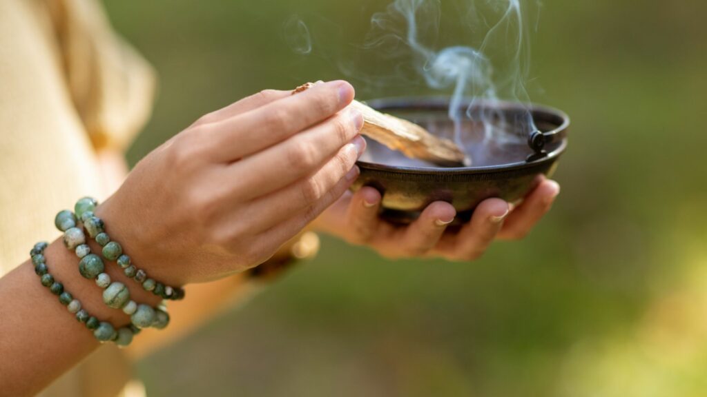 Imagem das mãos de uma mulher segurando em suas mãos um Palo Santo, realizando um ritual de limpeza energética.
