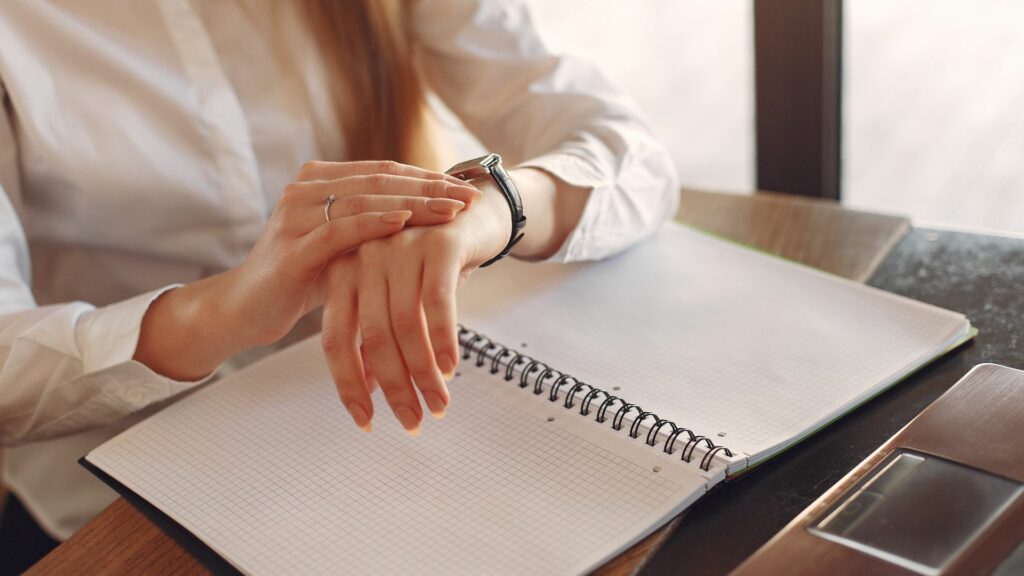 Imagem de uma mulher sentada, olhando no seu relógio de pulso, controlando o seu tempo. Sobre a mesa de trabalho, um caderno aberto.