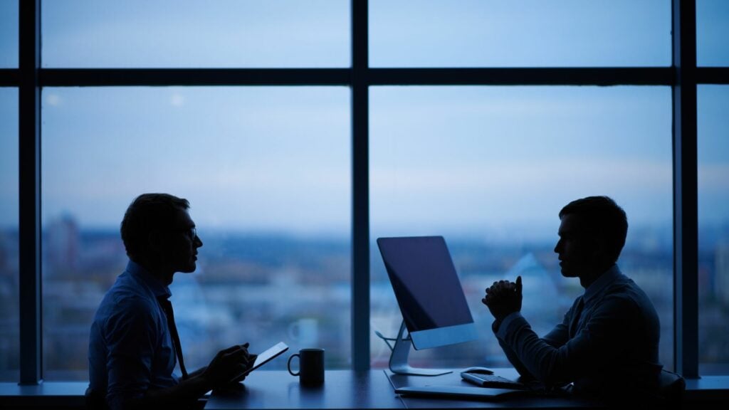 Imagem de dois homens conversando em uma sala de trabalho conversando sobre atividades profissionais.
