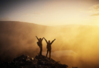 Imagem de uma montanha e o pôr do Sol. Em destaque um casal de mãos dadas e braços levantados, agradecendo a oportunidade de estarem vivos e vivendo a vida.