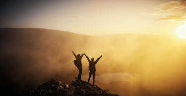 Imagem de uma montanha e o pôr do Sol. Em destaque um casal de mãos dadas e braços levantados, agradecendo a oportunidade de estarem vivos e vivendo a vida.